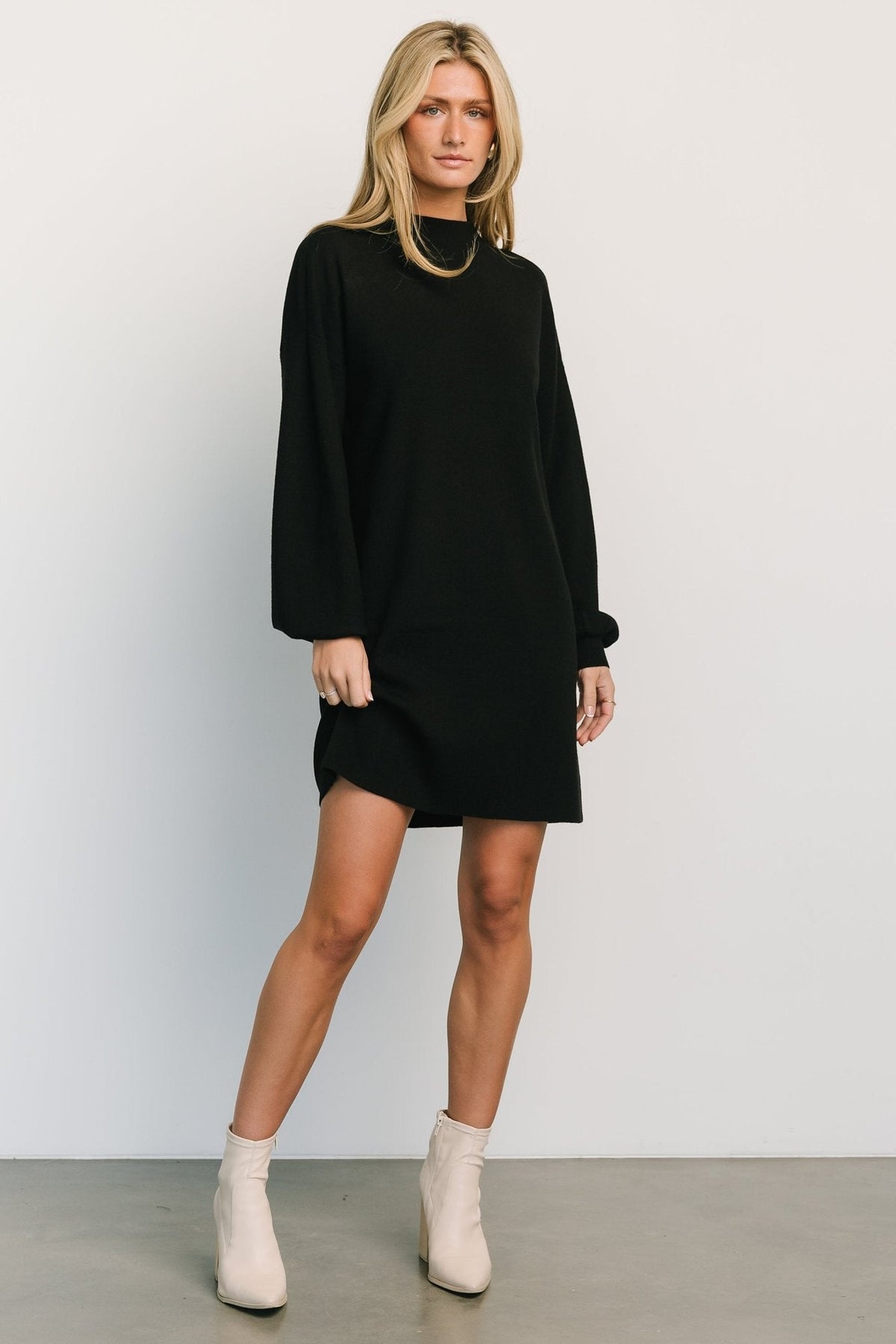 Front view of a short black sweater dress with mock neckline and long bubble sleeves on a model, styled with ankle boots and minimal accessories.